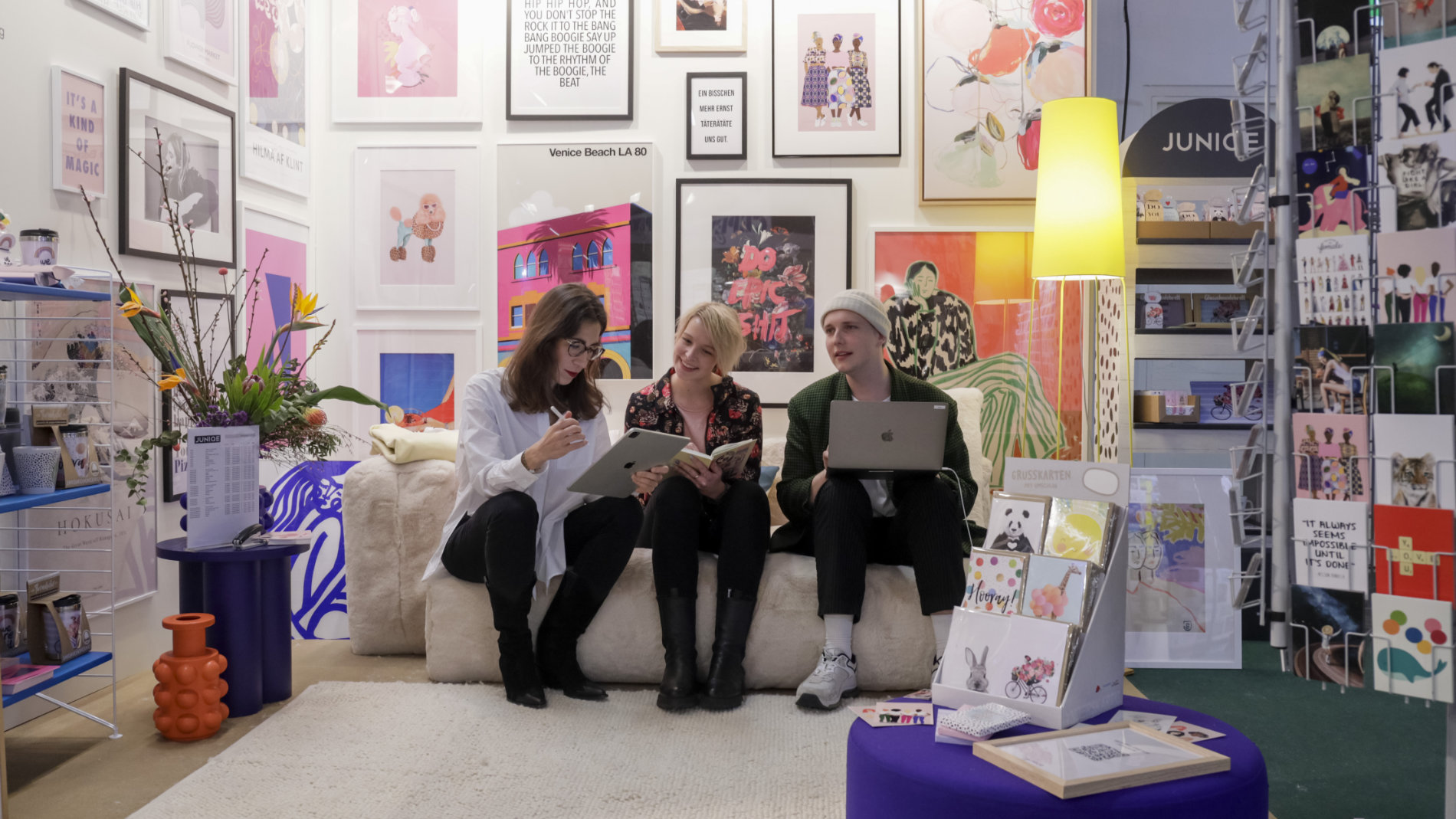 3 junge Leute sitzen an einem Ambiente Messestand mit Postern und Postkarten